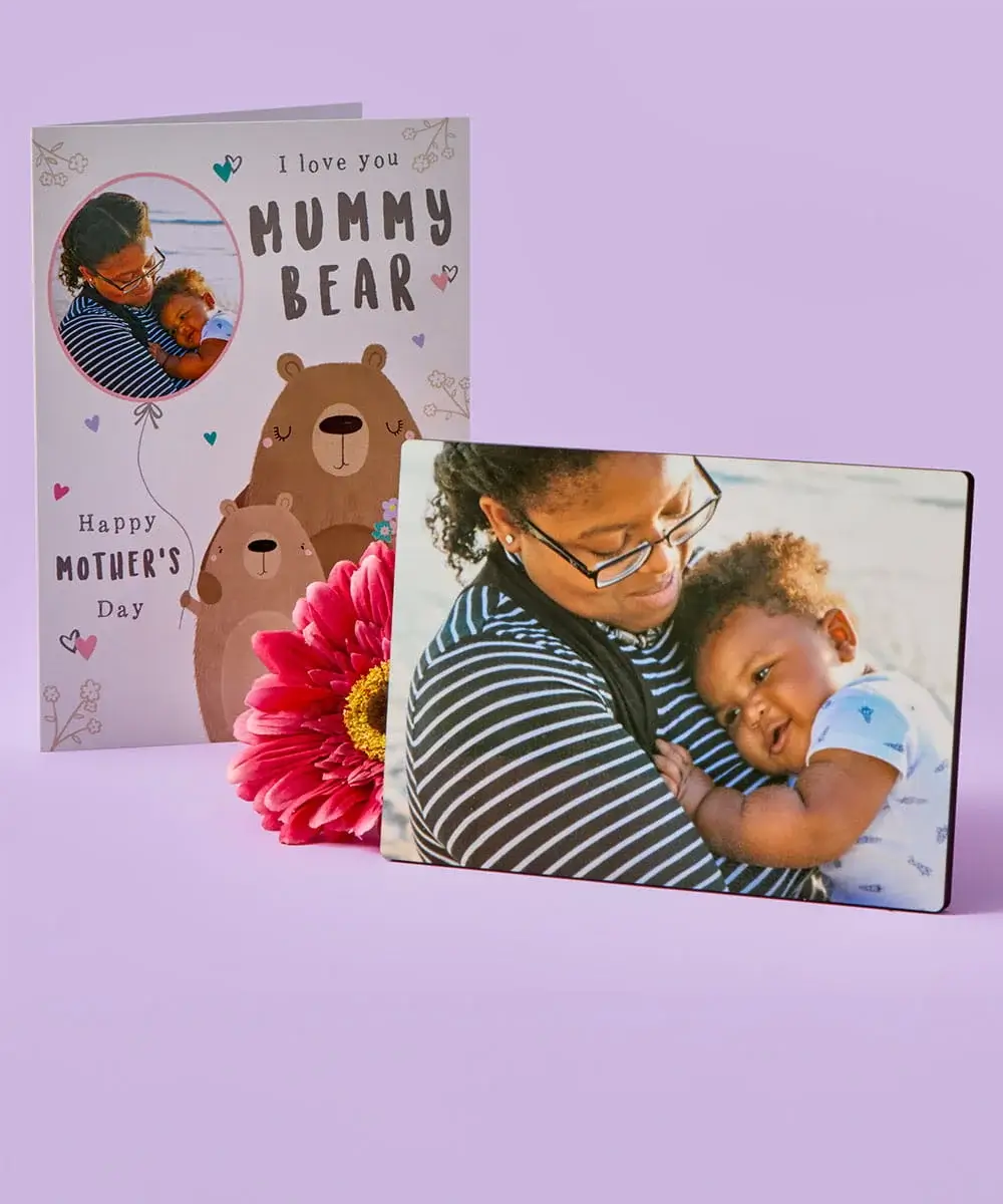 A Mother’s Day card with illustrated bears stands behind a photo plaque showing a parent holding a child, with a bright pink flower placed in front on a pastel purple background.